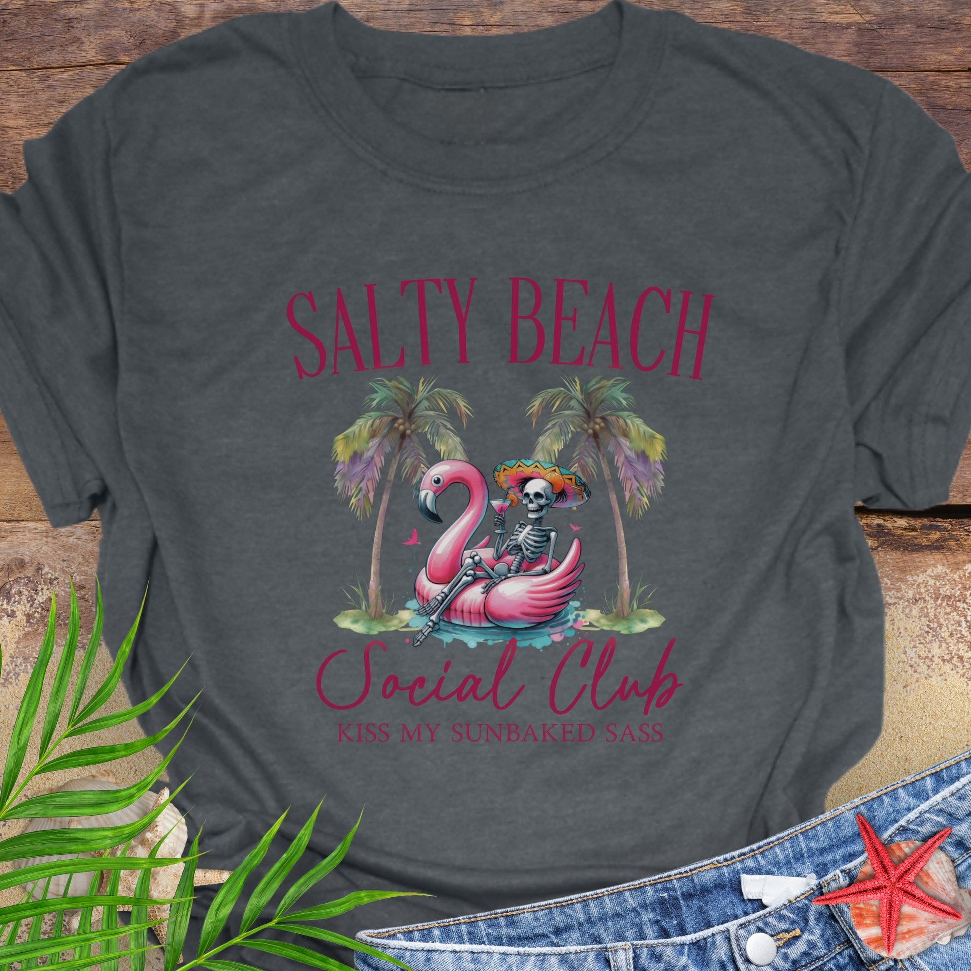Gray t-shirt with 'Salty Beach Social Club' design featuring a flamingo and skeleton, on a wooden surface with palm leaves and starfish.