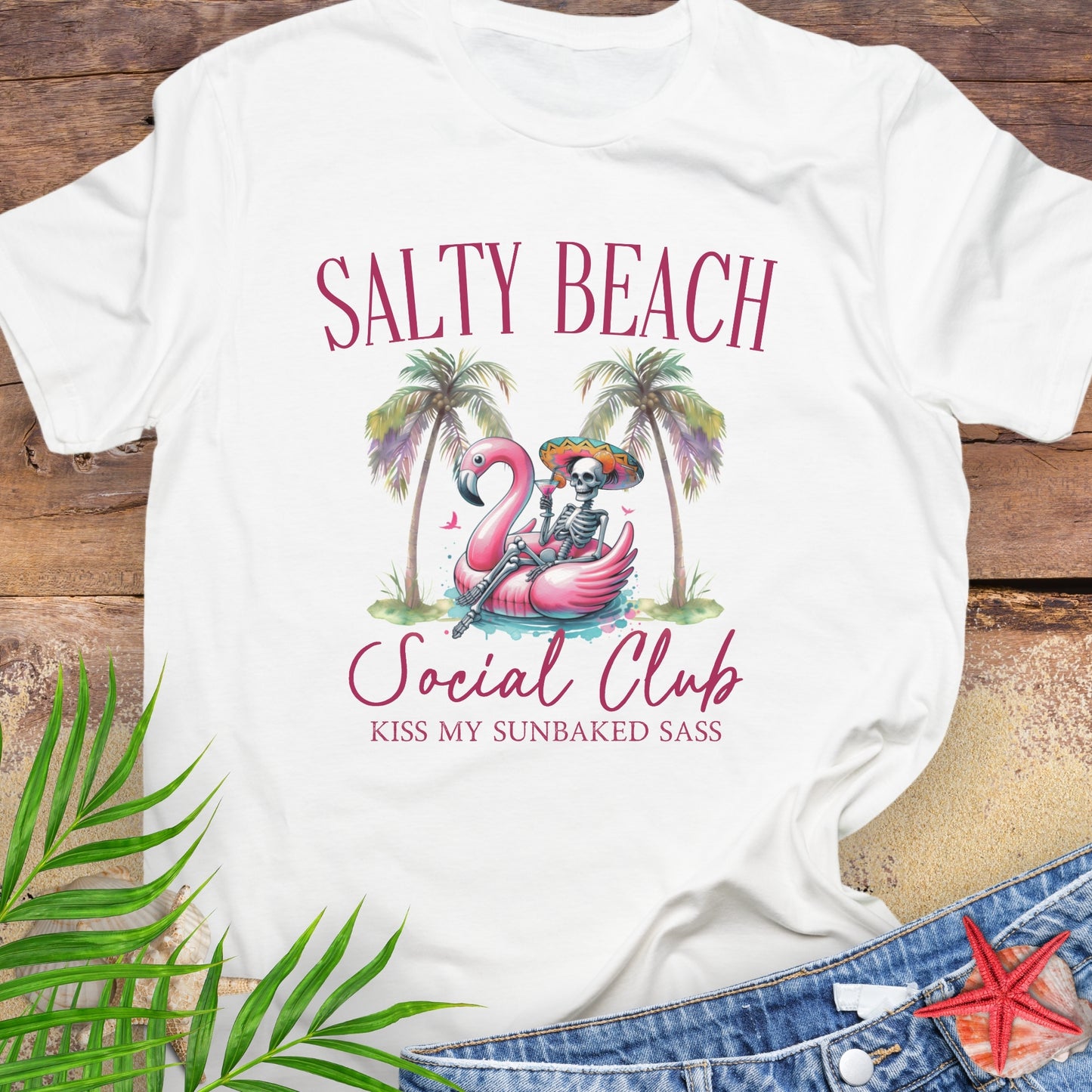 White t-shirt with 'Salty Beach Social Club' design featuring a flamingo and skeleton, on a wooden surface with palm leaves and denim jeans.