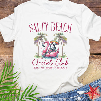 White t-shirt with 'Salty Beach Social Club' design featuring a flamingo and skeleton, on a wooden surface with palm leaves and denim jeans.
