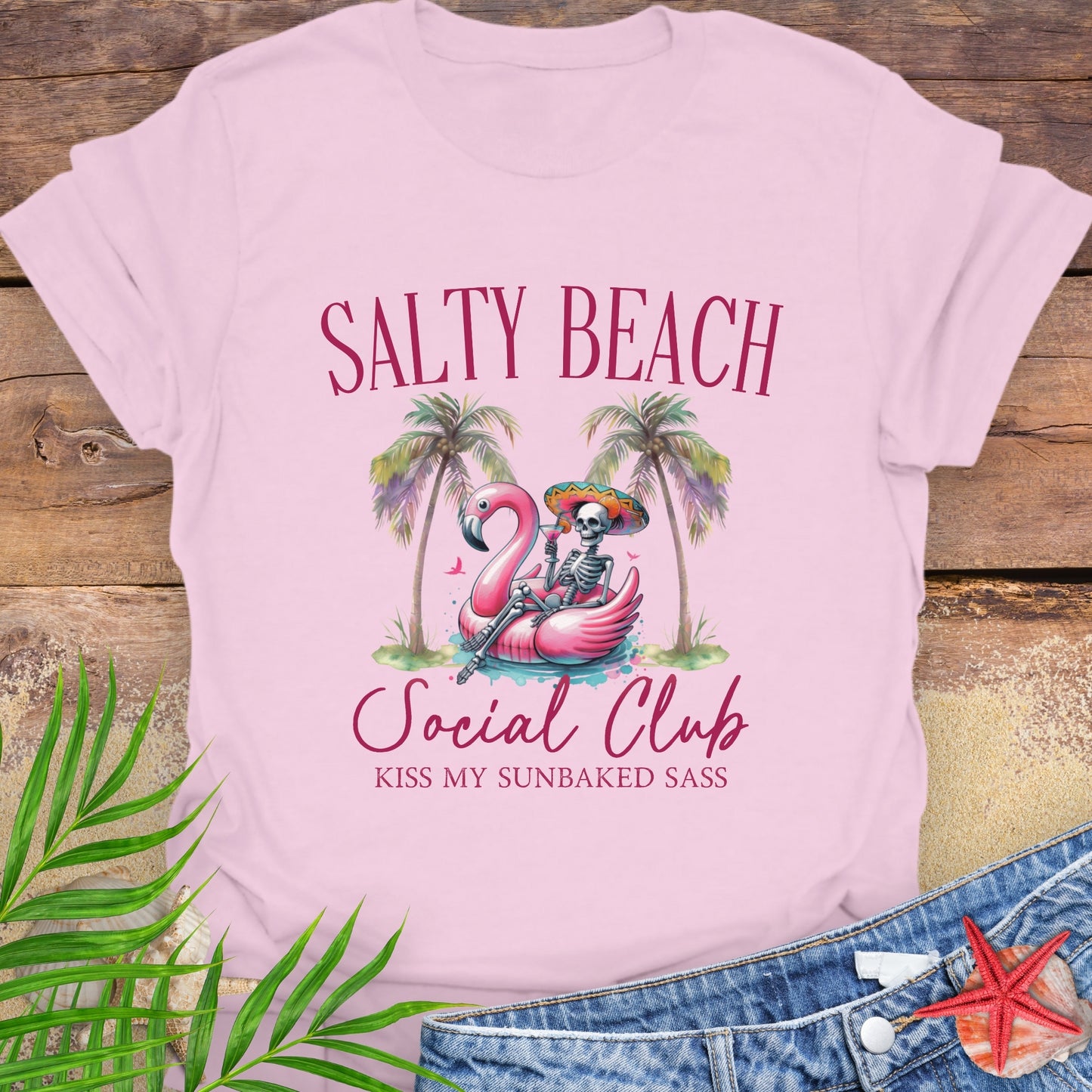 Pink t-shirt with 'Salty Beach Social Club' design featuring a skeleton on a flamingo, palm trees, and beach text.