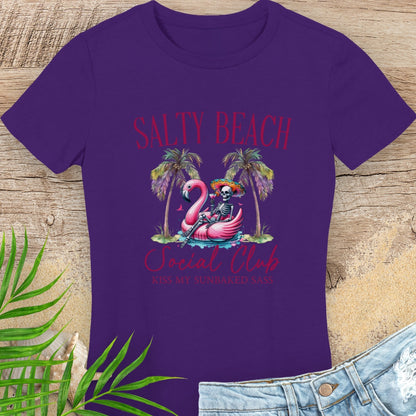 Purple t-shirt with 'Salty Beach Social Club' design featuring a skeleton and flamingo, on a wooden surface with palm leaves and denim jeans.
