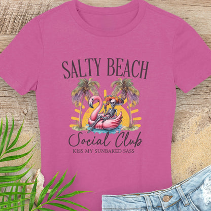 Pink t-shirt with 'Salty Beach Social Club' design featuring a flamingo and skeleton, on a wooden surface with beach elements.