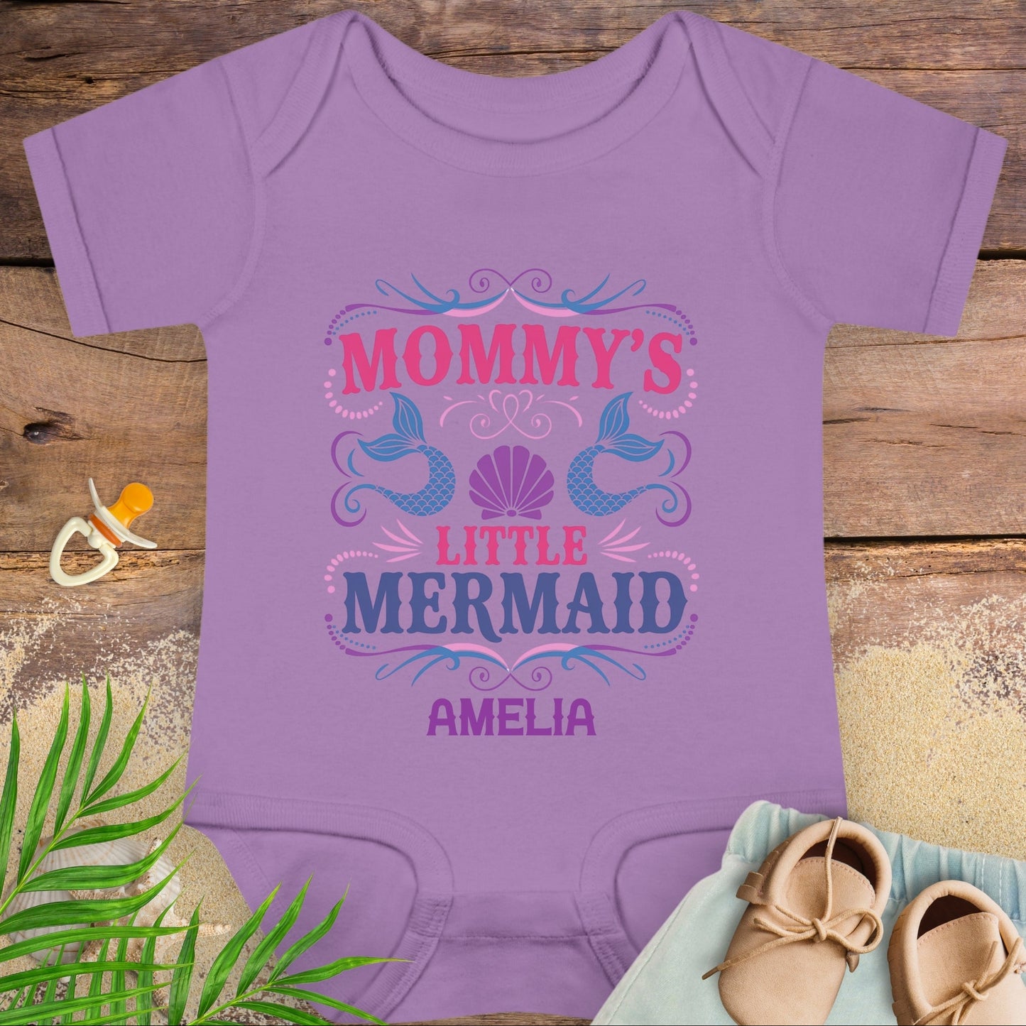 Purple baby onesie with 'Mommy's Little Mermaid' design on a wooden surface with a pacifier and shoes.