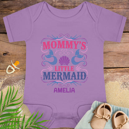 Purple baby onesie with 'Mommy's Little Mermaid' design on a wooden surface with a pacifier and shoes.