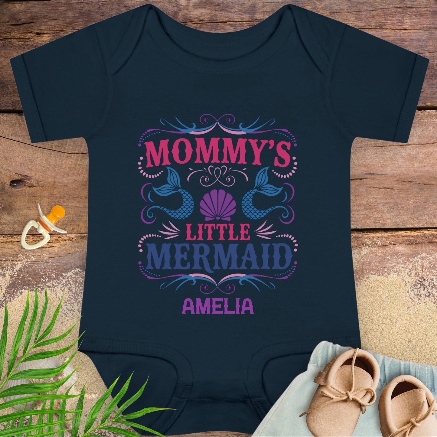 Navy blue baby onesie with 'Mommy's Little Mermaid Amelia' text on a wooden surface with a pacifier and shoes.