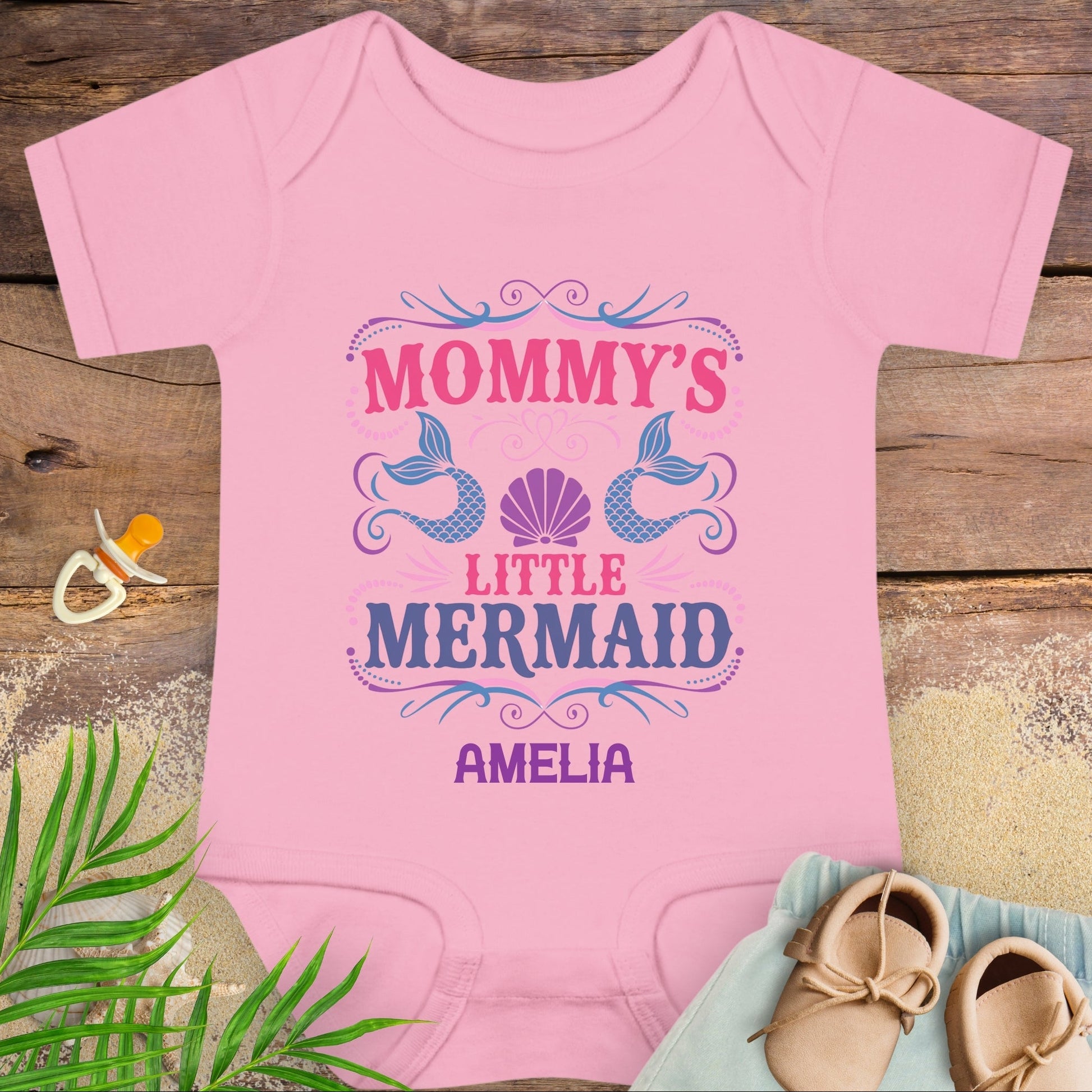 Pink baby onesie with 'Mommy's Little Mermaid' design on a wooden surface with baby items.