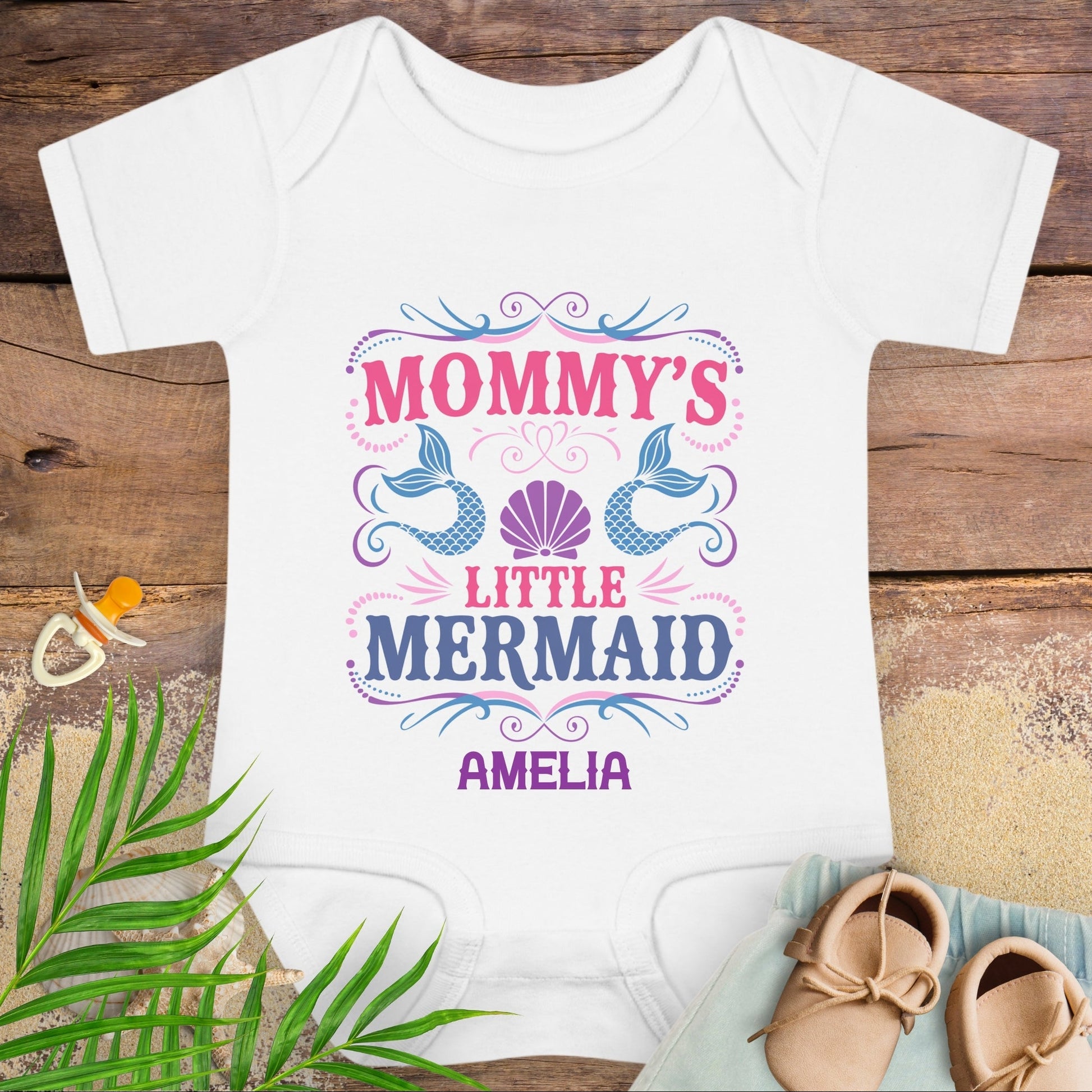 White baby onesie with 'Mommy's Little Mermaid Amelia' text on a wooden surface with a pacifier and shoes.