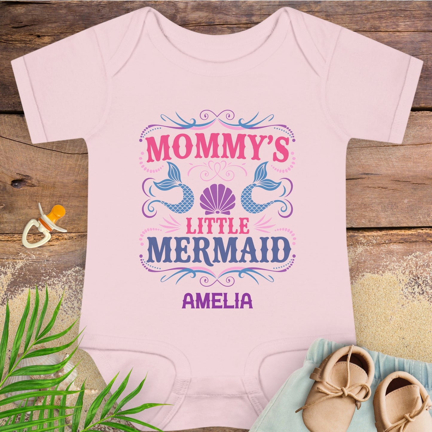 Pink baby onesie with 'Mommy's Little Mermaid' design on a wooden surface with shoes and a pacifier.