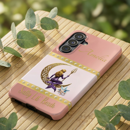 Phone case with a design of a mermaid and crescent moon on a pink background, placed on a wooden surface.