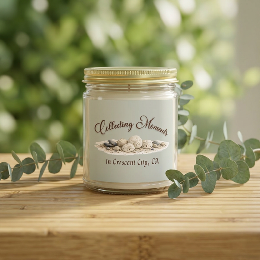 A glass jar candle with a gold metal lid sits on a wooden surface, surrounded by eucalyptus leaves. The candle has a sage green label that reads 'Collecting Moments in Crescent City, CA' with an illustration of seashells.