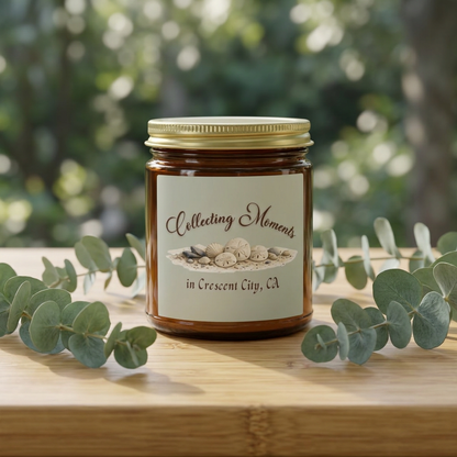 An amber glass jar candle with a gold metal lid sits on a wooden surface, surrounded by eucalyptus leaves. The candle has a sage green label that reads 'Collecting Moments in Crescent City, CA' with an illustration of seashells.