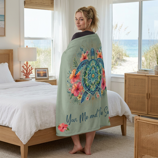 A woman stands in a bedroom wrapped in a sage green ultra-soft plush blanket featuring a vibrant mandala sea turtle surrounded by tropical hibiscus flowers with the phrase "You, Me and the Sea" on a sage-green background.