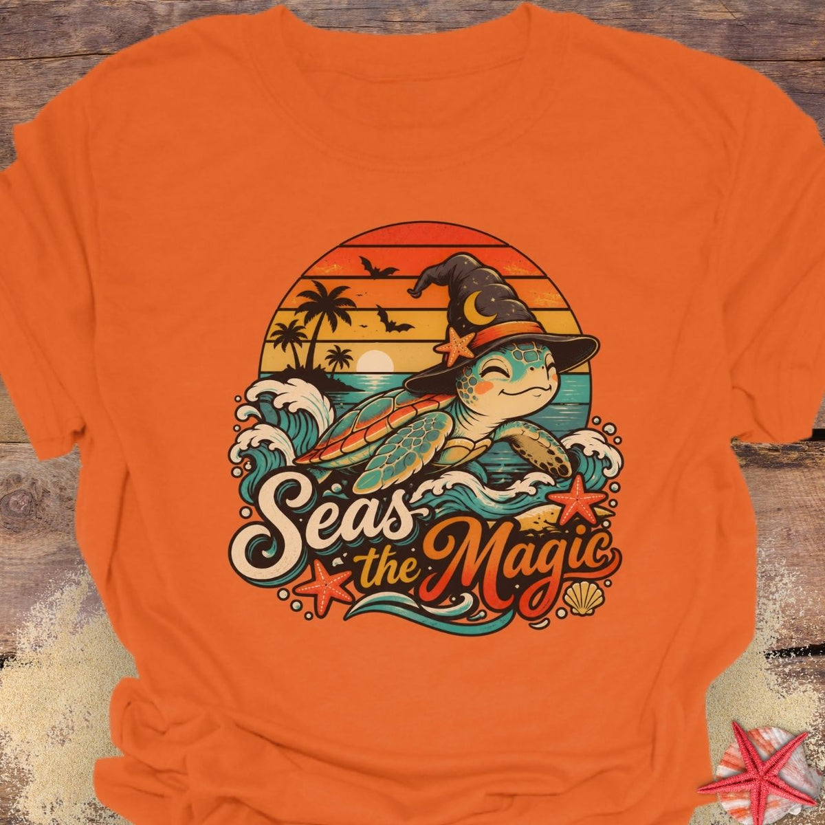 Orange unisex t-shirt with “Seas the Magic” text, a sea turtle wearing a witch hat, ocean waves, sunset, palm trees, bats, and starfish.