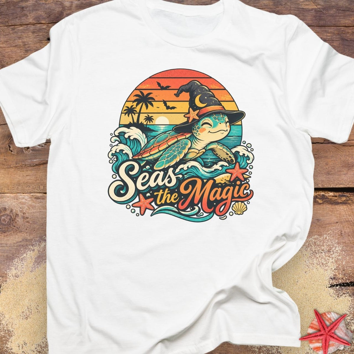 White unisex t-shirt with “Seas the Magic” text, a sea turtle wearing a witch hat, ocean waves, sunset, palm trees, bats, and starfish.