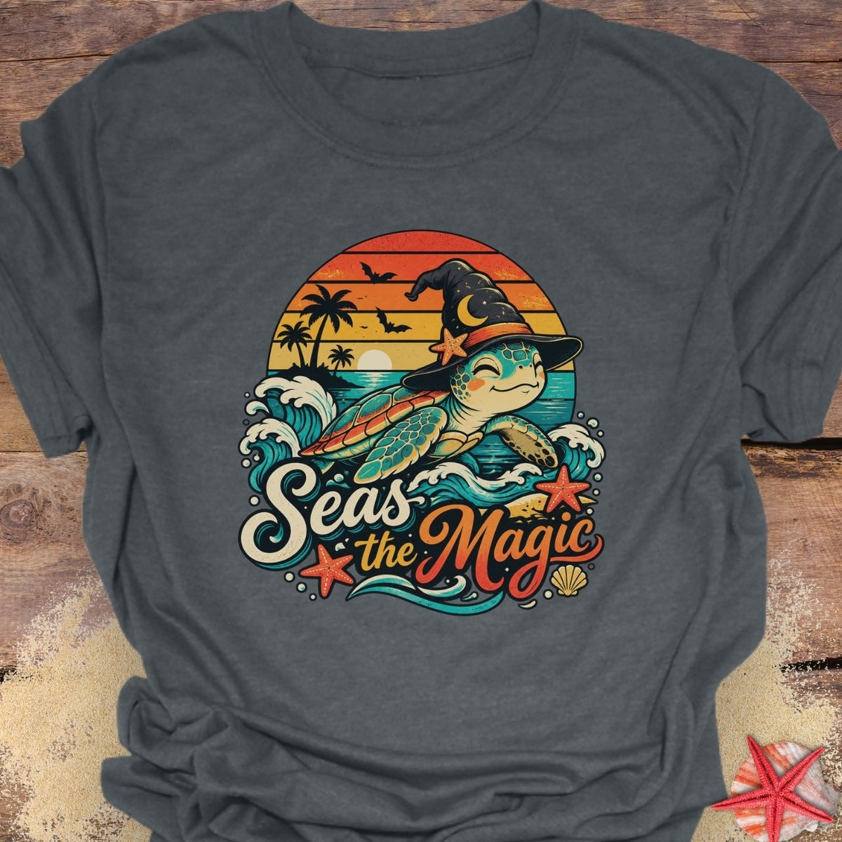 dark heather unisex t-shirt with “Seas the Magic” text, a sea turtle wearing a witch hat, ocean waves, sunset, palm trees, bats, and starfish.