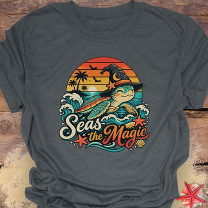 dark heather unisex t-shirt with “Seas the Magic” text, a sea turtle wearing a witch hat, ocean waves, sunset, palm trees, bats, and starfish.