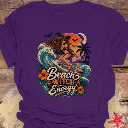 Purple unisex t-shirt with “Beach Witch Energy” text, a witchy surfer girl, ocean wave, sunset, crescent moon, bats, palm trees, and tropical flowers.
