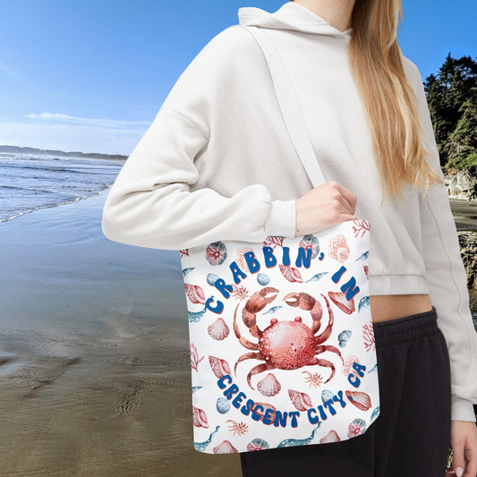 White 13" x 13" Tote bag featuring a crab surrounded by seashells and underwater life in blue and soft red tones with the phrase "Crabbin' in Crescent City, CA.
