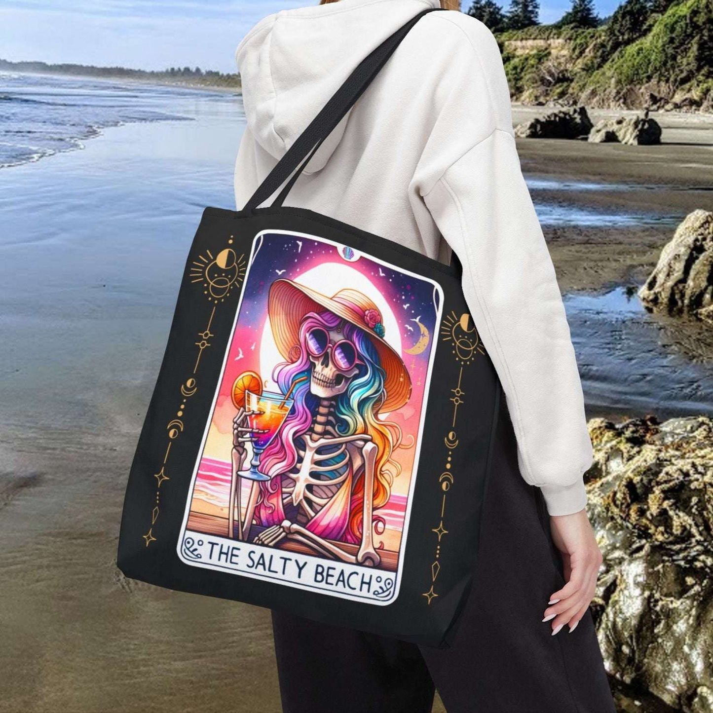 Person holding a tote bag with a colorful skeleton woman holding a cocktail with a colorful beach sunset on a tarot card design with text The Salty Beach.