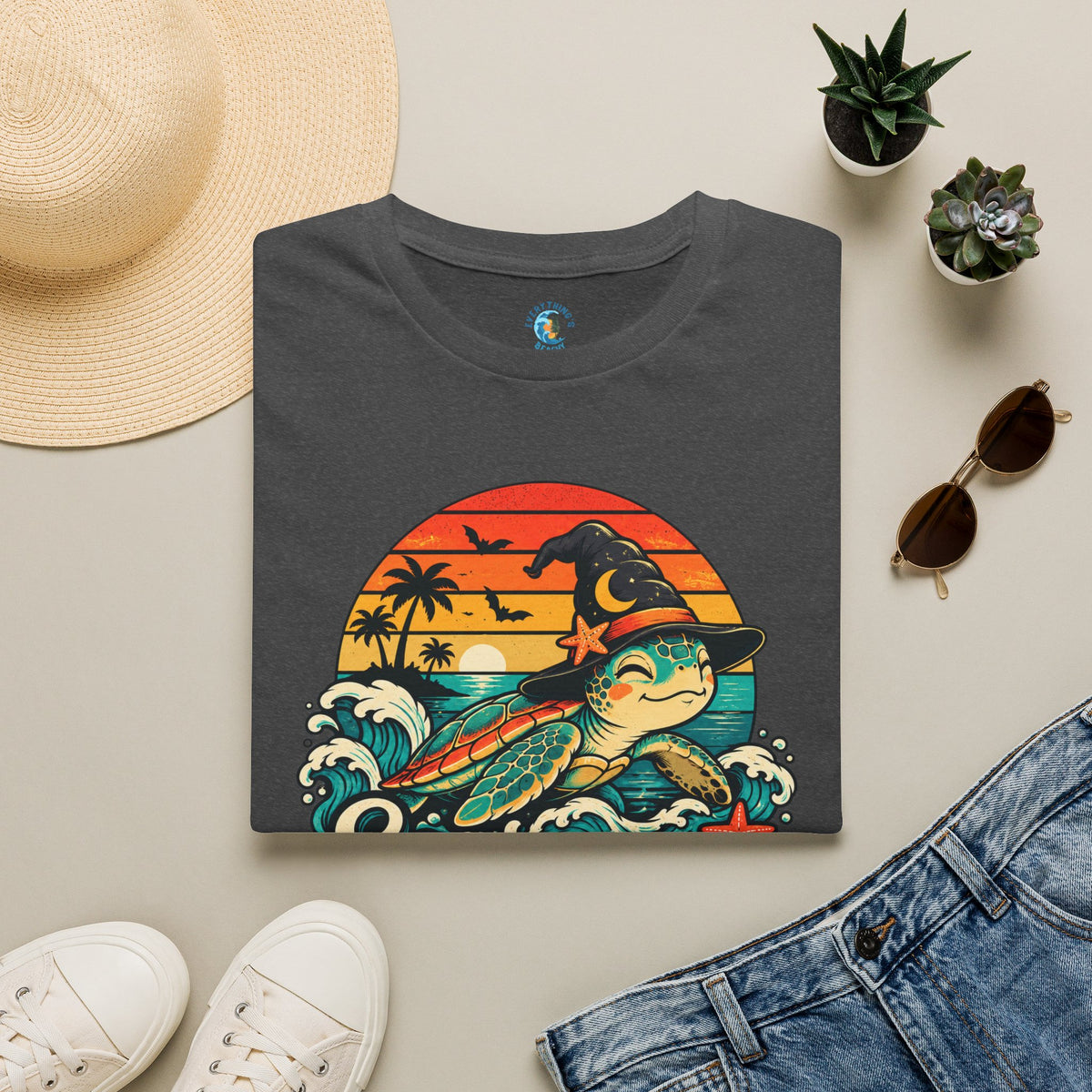Folded dark heather gray unisex t-shirt with “Seas the Magic” text, a sea turtle wearing a witch hat, ocean waves, sunset, palm trees, bats, and starfish.
