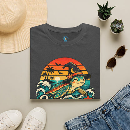 Folded dark heather gray unisex t-shirt with “Seas the Magic” text, a sea turtle wearing a witch hat, ocean waves, sunset, palm trees, bats, and starfish.