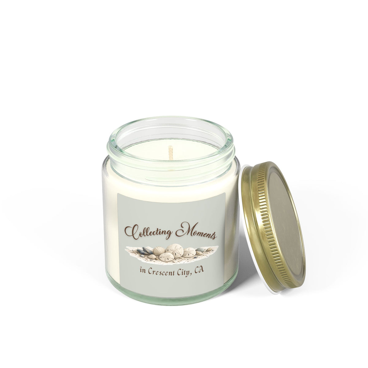 A glass jar candle with a gold metal lid sits on a wooden surface, surrounded by eucalyptus leaves. The candle has a sage green label that reads 'Collecting Moments in Crescent City, CA' with an illustration of seashells.