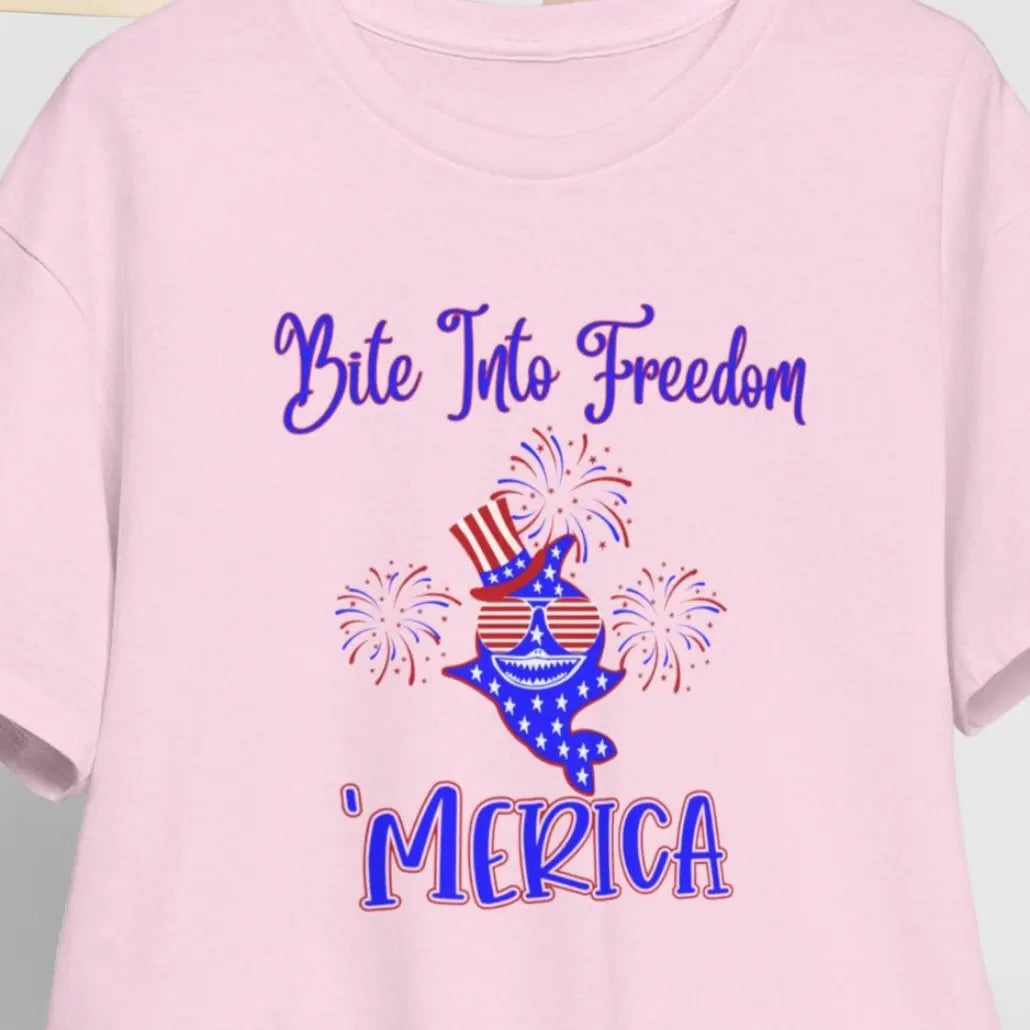 A pink t-shirt on a hanger with a patriotic shark with red and white striped glasses, a patriotic top hat with fireworks around that says Bite Into Freedom "merica.