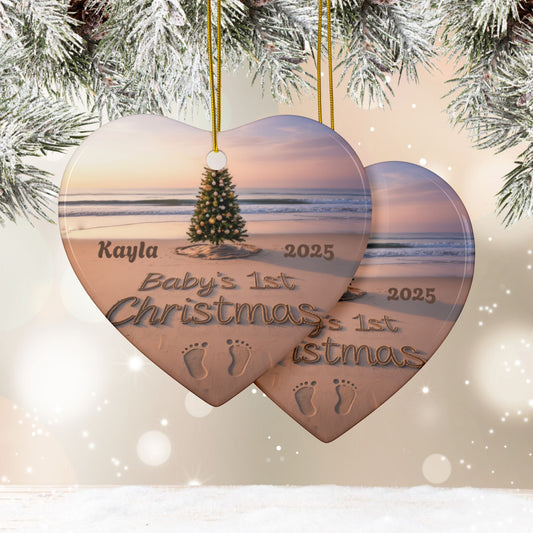 Heart-shaped Christmas ornament with beach scene and 'Baby's 1st Christmas' text, hanging against a snowy branch background.