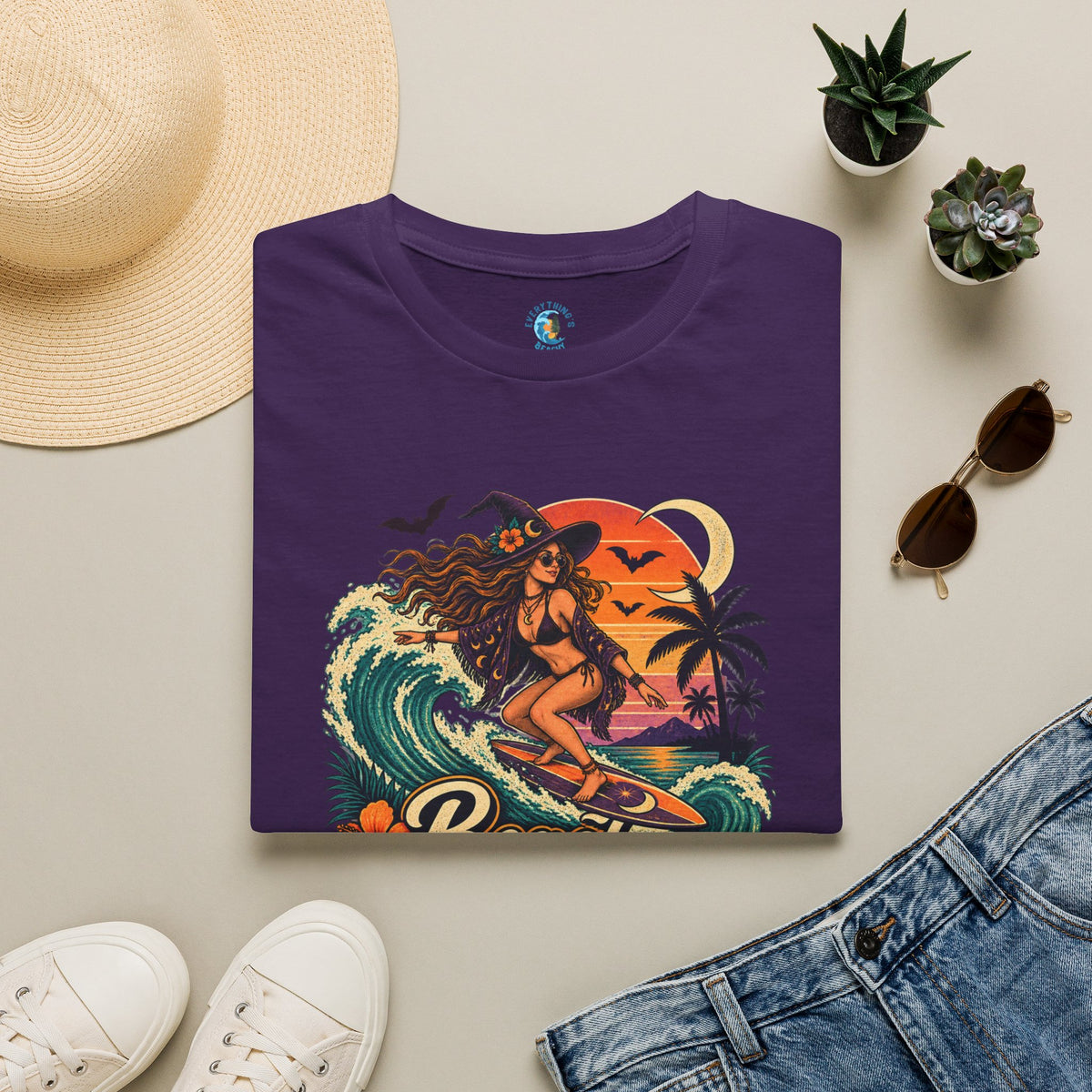Folded purple unisex t-shirt with “Beach Witch Energy” text, a witchy surfer girl, ocean wave, sunset, crescent moon, bats, palm trees, and tropical flowers.
