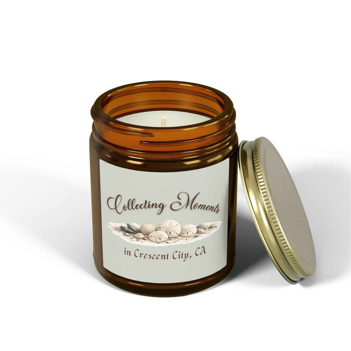 An amber glass jar candle with a gold metal lid sits on a wooden surface, surrounded by eucalyptus leaves. The candle has a sage green label that reads 'Collecting Moments in Crescent City, CA' with an illustration of seashells.