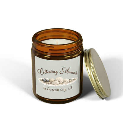 An amber glass jar candle with a gold metal lid sits on a wooden surface, surrounded by eucalyptus leaves. The candle has a sage green label that reads 'Collecting Moments in Crescent City, CA' with an illustration of seashells.