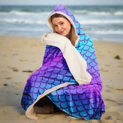 Person sitting on the beach wearing a hooded blanket with mermaid scales in purple and turquoise with a picture of a beautiful mermaid with the words mermaid vibes below