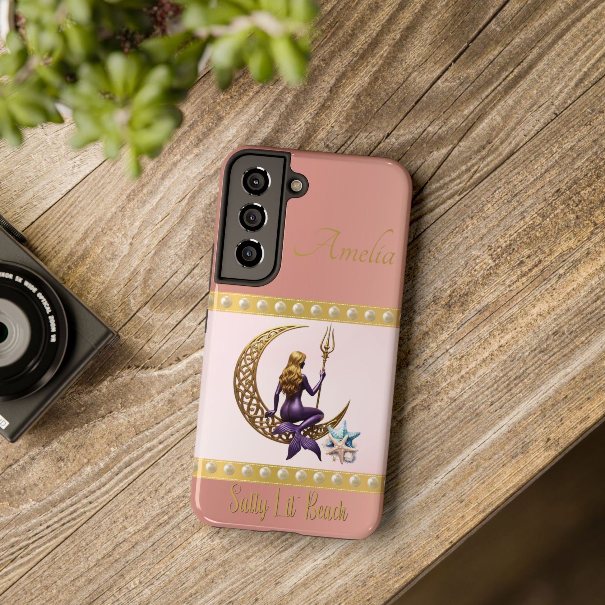 Phone case with mermaid design on a wooden surface next to a camera and plant