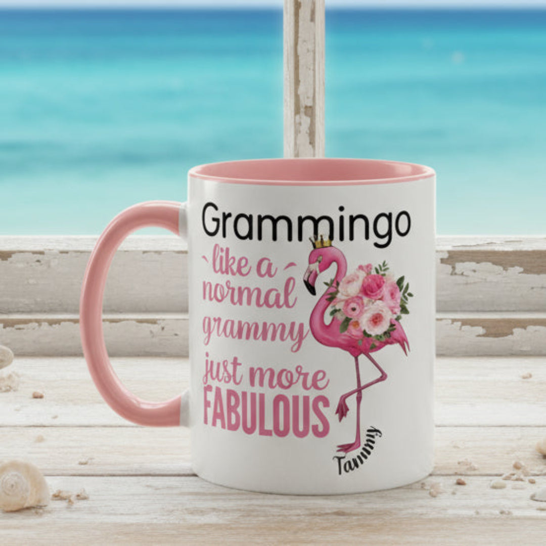 Ceramic coffee mug featuring a flamingo adorned with flowers and the text “Grammingo like a normal Grammy just more fabulous,” available with white, pink, or black handle options.