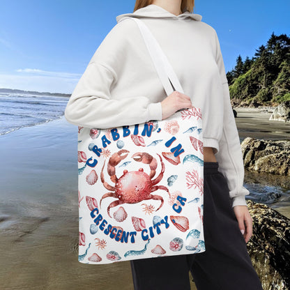 White 18" x 18" Tote bag featuring a crab surrounded by seashells and underwater life in blue and soft red tones with the phrase "Crabbin' in Crescent City, CA.