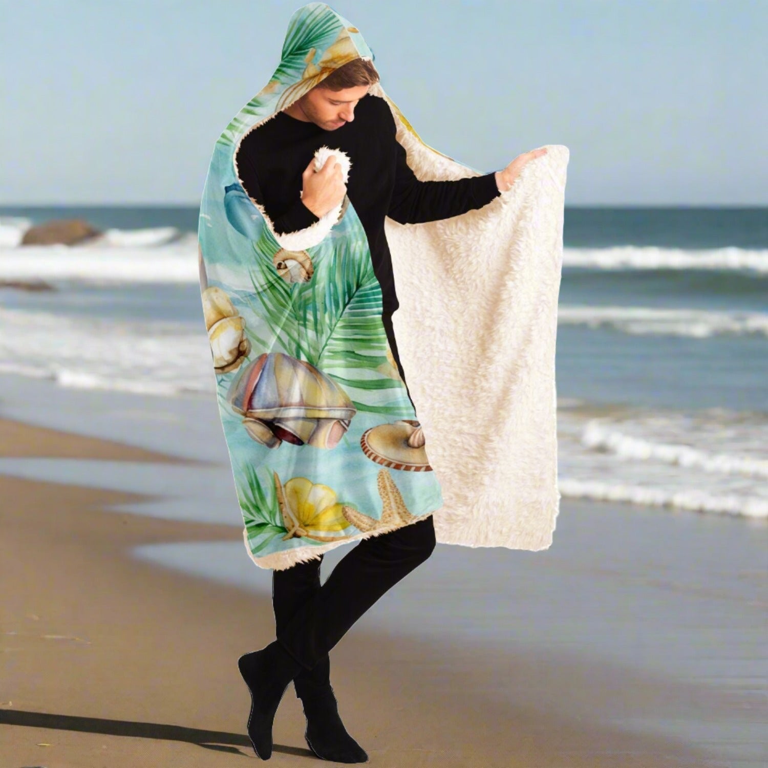 Person standing on a beach holding open a hooded blanket with a light aqua background with seashells and leaves with blue text that says Chill at the Beach showing the soft inside lining.