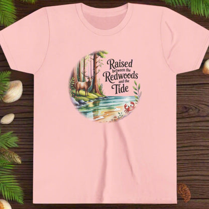 Pink t-shirt with nature-themed graphic with elk among redwoods and stream with a crab and text Raised between the Redwoods and the Tide.