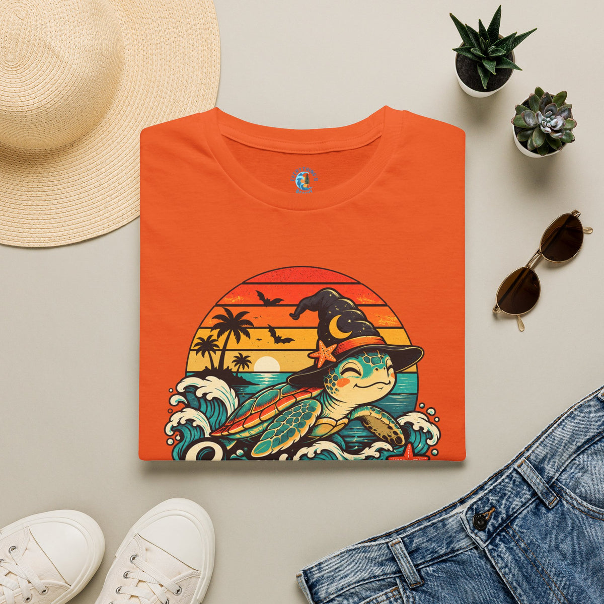 Folded Orange unisex t-shirt with “Seas the Magic” text, a sea turtle wearing a witch hat, ocean waves, sunset, palm trees, bats, and starfish.