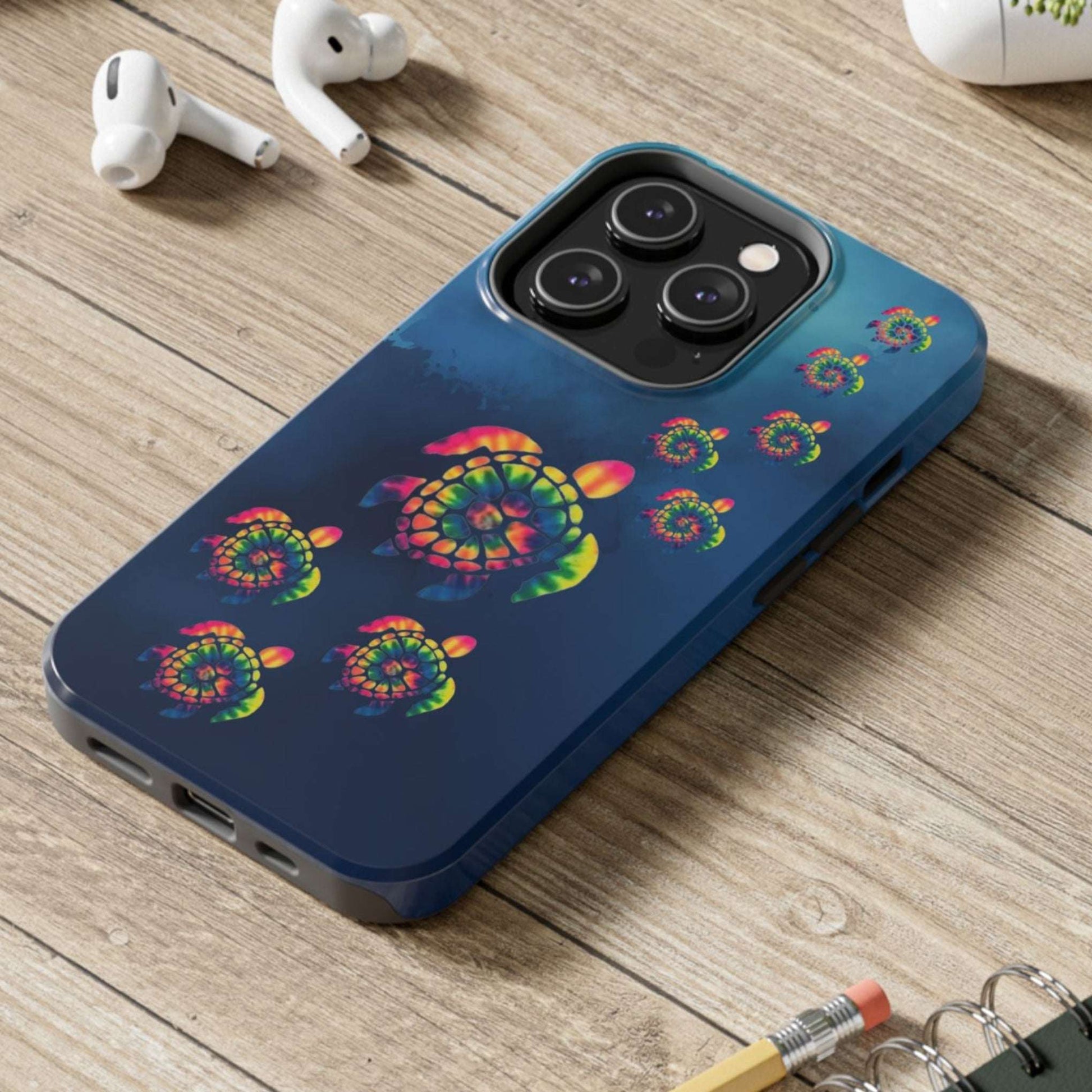 Phone case with colorful turtle design on a wooden surface with laptop, earbuds, and notebook.