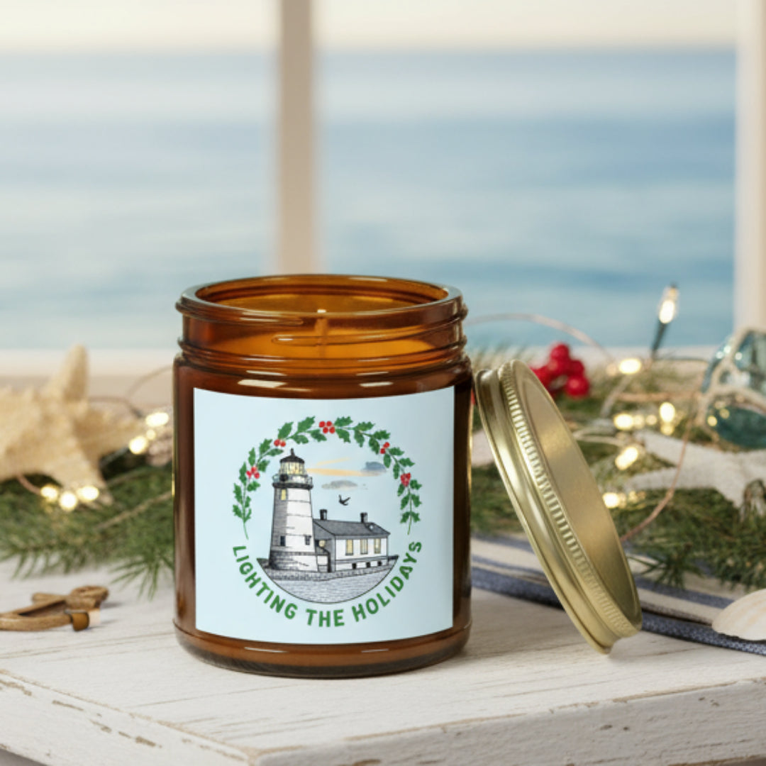 Candle in an amber jar with a decorative label featuring a lighthouse and holly wreath on a white background.