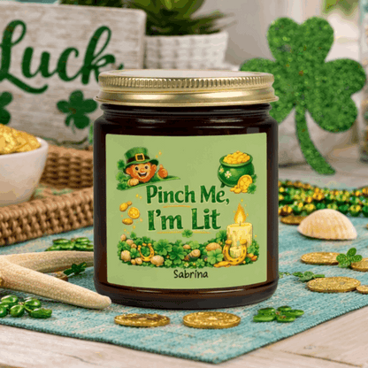 Amber candle jar with a lit candle, green label, and gold lid on with St. Patrick's Day Design and text Pinch Me, I'm Lit that can be personalized.