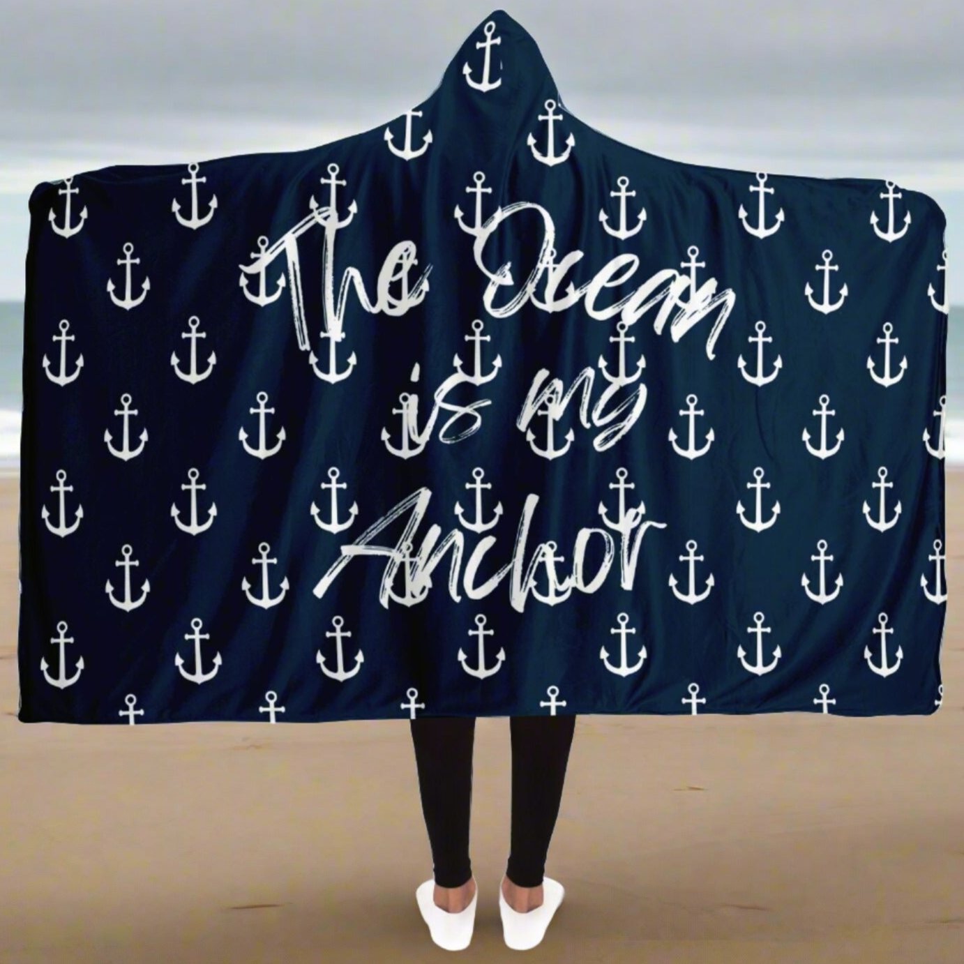 Person standing on the beach in an open dark blue hooded blanket with white anchors and white writing that says The Ocean is my Anchor