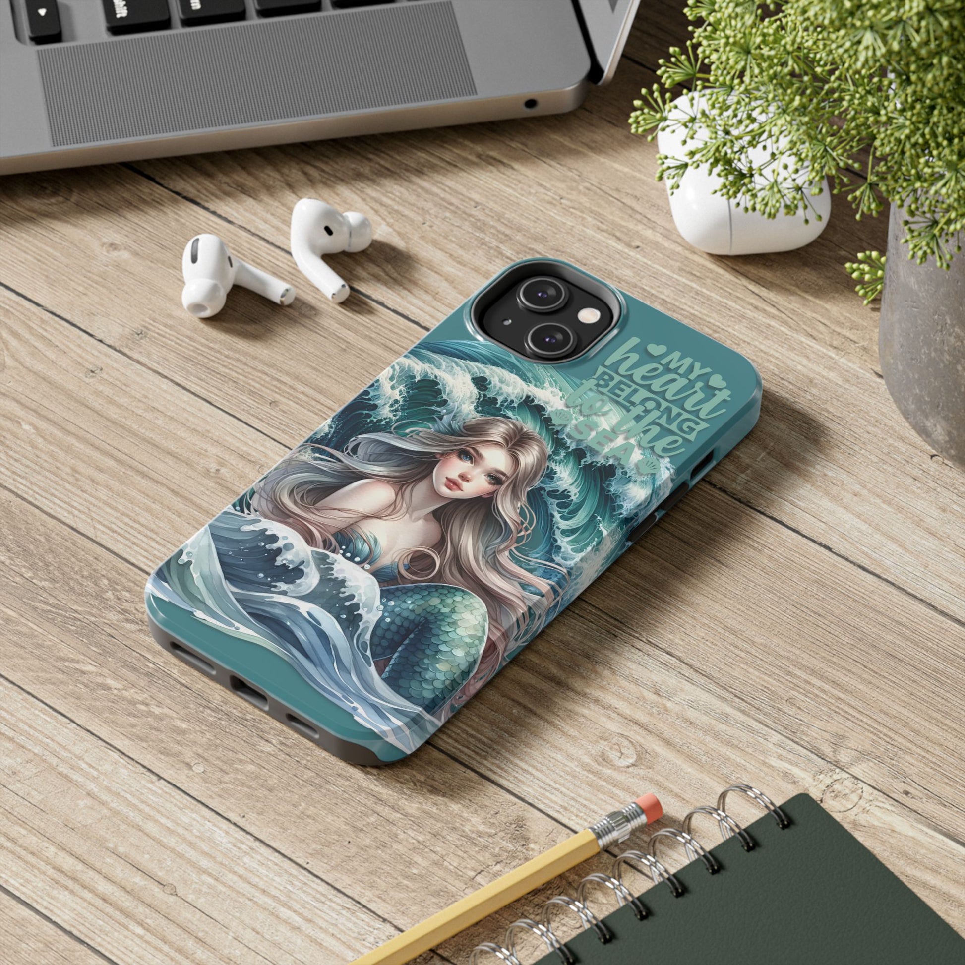 Phone Case with Flowy mermaid with ocean wave behind and text My Heart Belongs to the Sea laying on desk.