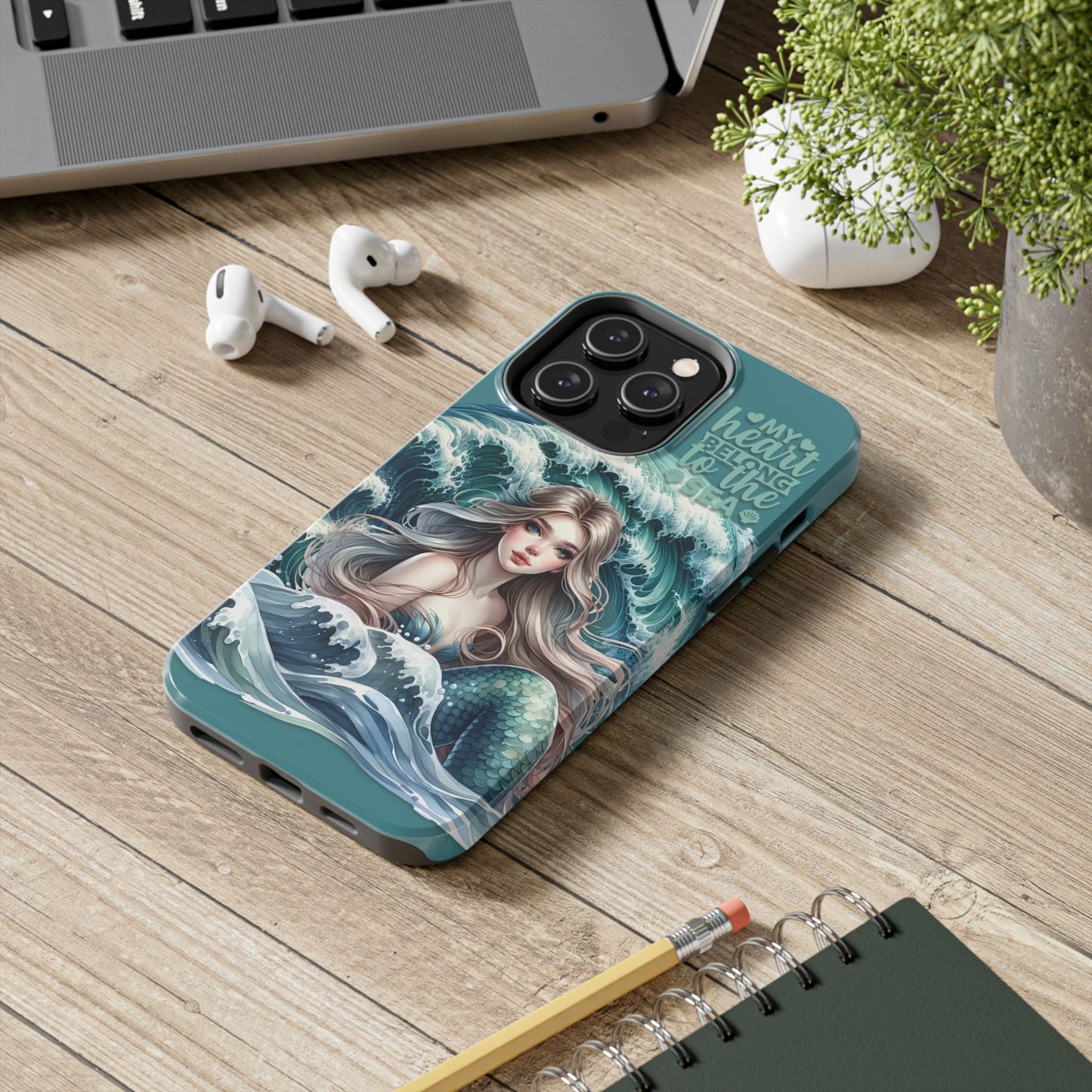 Phone Case with Flowy mermaid with ocean wave behind and text My Heart Belongs to the Sea laying on desk.