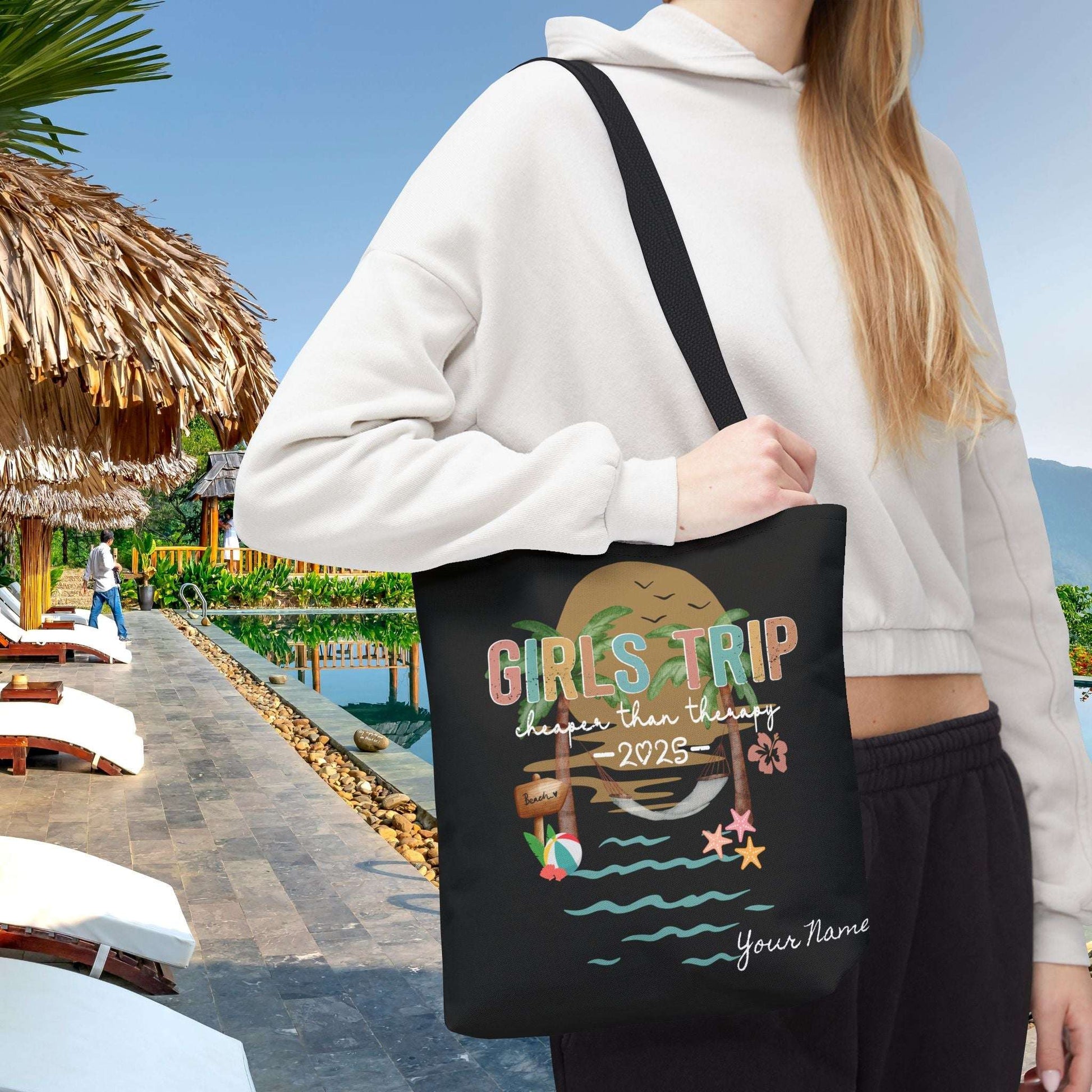 Person holding a 13" x 13" black tote bag with 'Girls Trip' beach design that can be personalized.