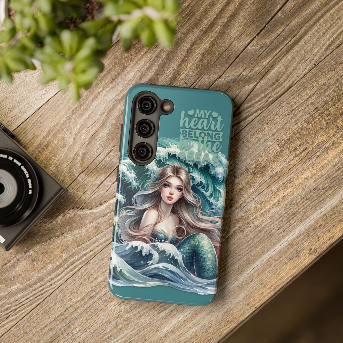 Phone Case with Flowy mermaid with ocean wave behind and text My Heart Belongs to the Sea laying on desk.