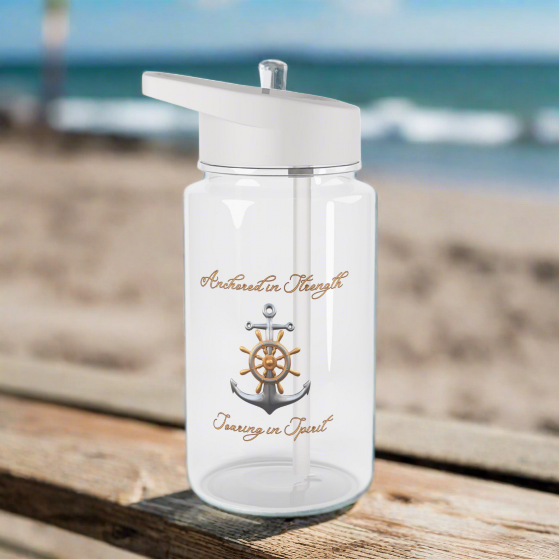 Clear Water bottle with gray lid and straw with an anchor that says anchored in strength and soaring in spirit sitting on a picnic table at the beach