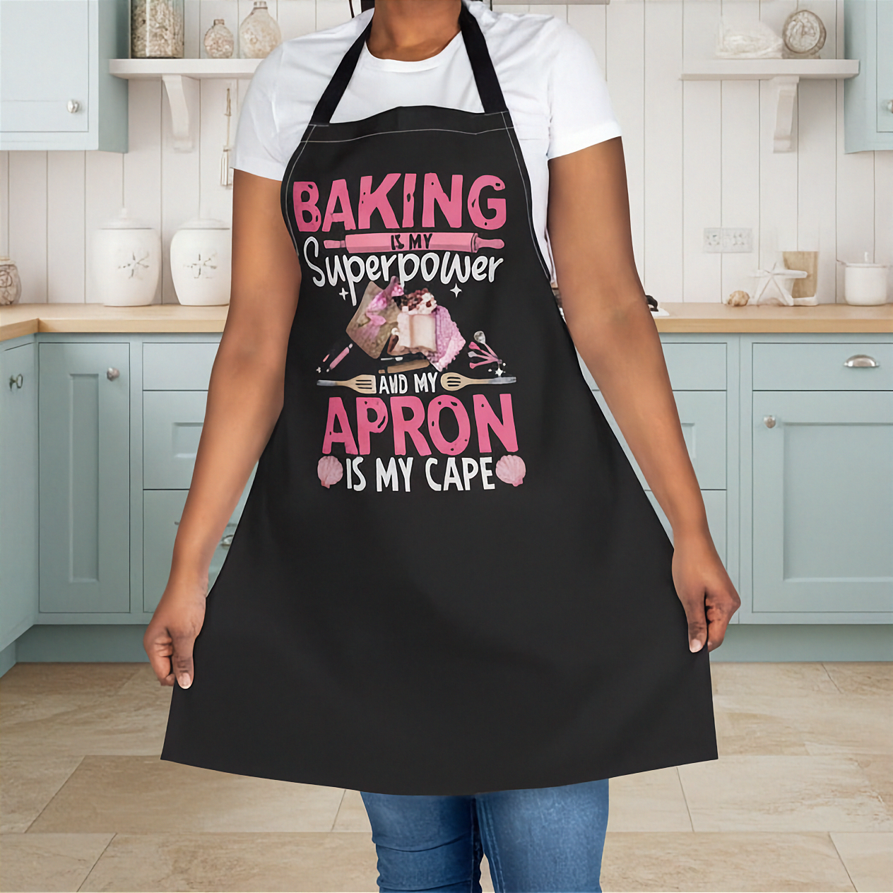 A person wearing a black kitchen apron with pink and white text that reads 'BAKING IS MY Superpower AND MY APRON IS MY CAPE' along with a baking-themed graphic, standing in a kitchen with light blue cabinets.