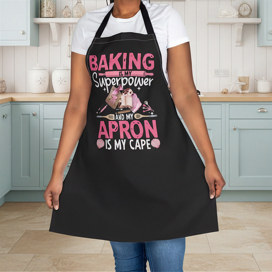 A person wearing a black kitchen apron with pink and white text that reads 'BAKING IS MY Superpower AND MY APRON IS MY CAPE' along with a baking-themed graphic, standing in a kitchen with light blue cabinets.