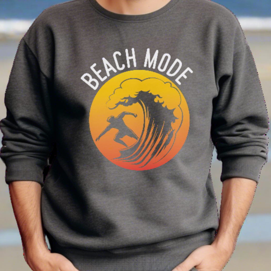 A man standing on the beach in a dark heather crewneck sweatshirt with a Person surfing a wave with a yellow and orange sunset background that says Beach Mode.